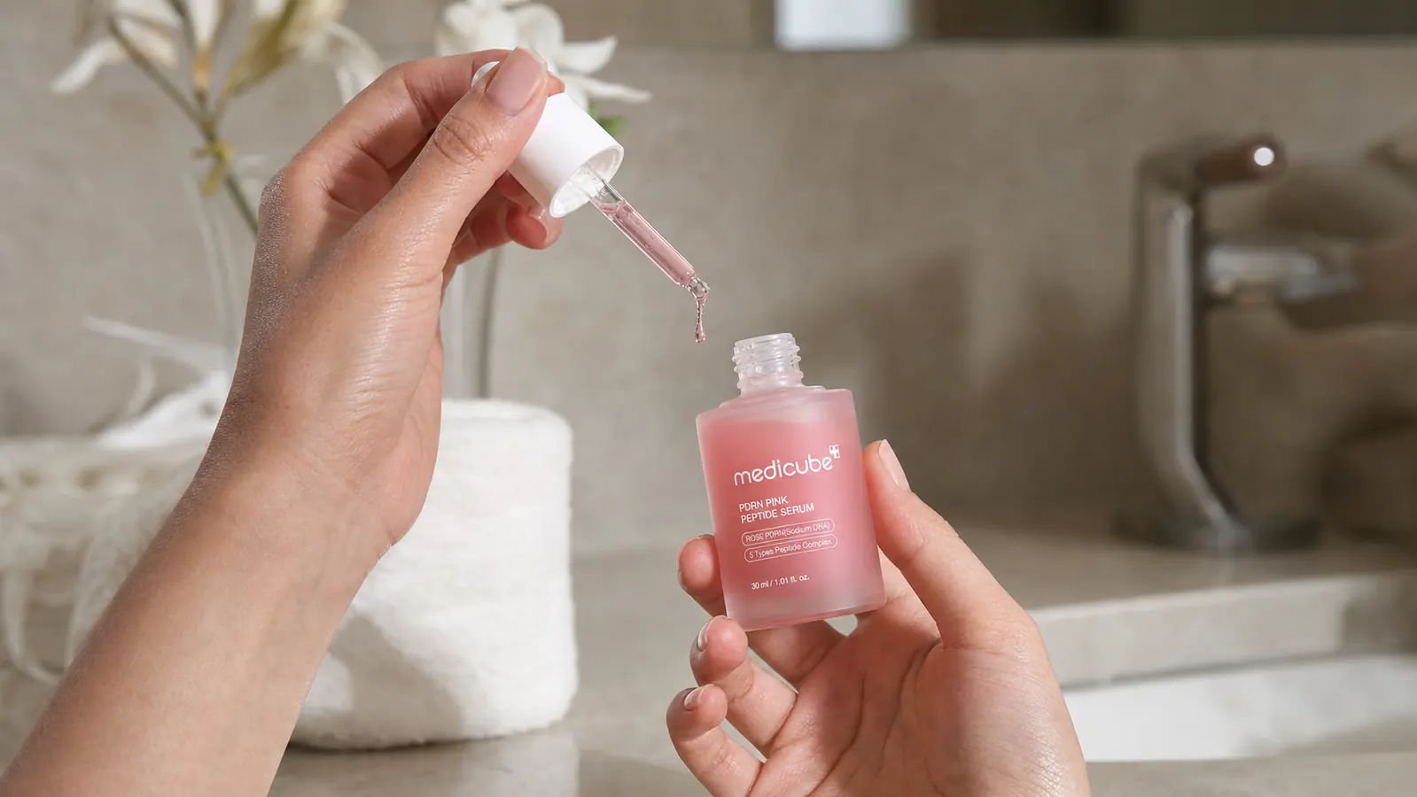 Close-up of a dropper dispensing pink serum onto fingertips, with the medicube bottle visible in the background