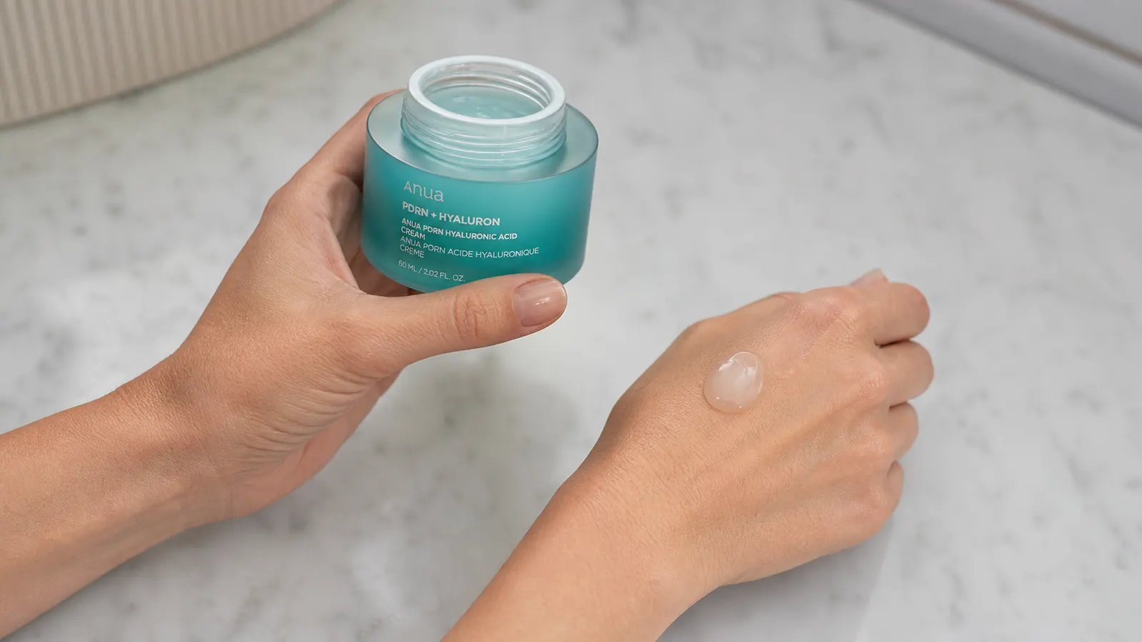 Anua PDRN Hyaluronic Acid 100 Moisturizing Cream jar with a small amount of cream on the back of a hand showing the lightweight gel-cream texture