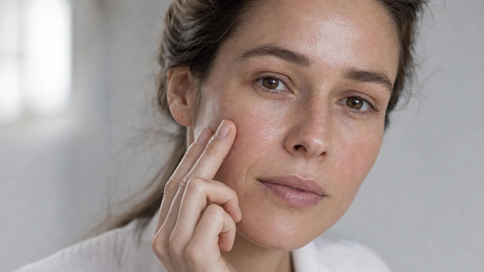 Close-up of a woman touching her cheek with fingertips, skin appearing dewy and smooth in natural window light