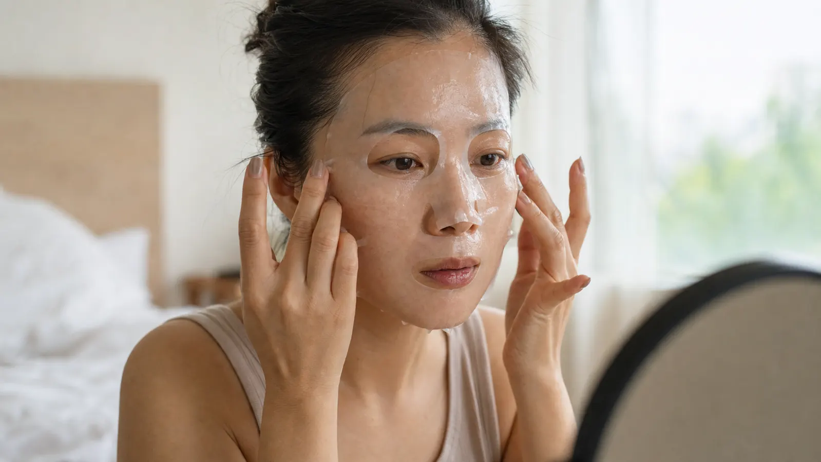 Woman with glowing skin in soft morning light holding a hydrogel mask sheet next to her cheek, eyes closed, relaxed expression