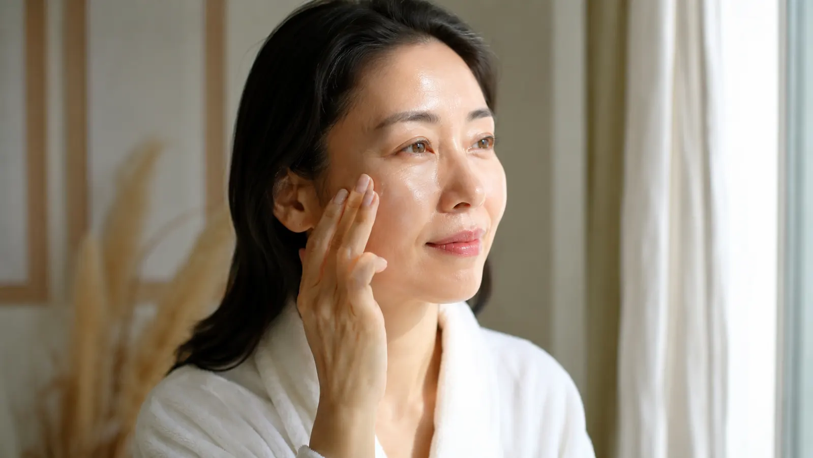 Woman with clear glowing skin applying serum to her cheek in morning light near a window